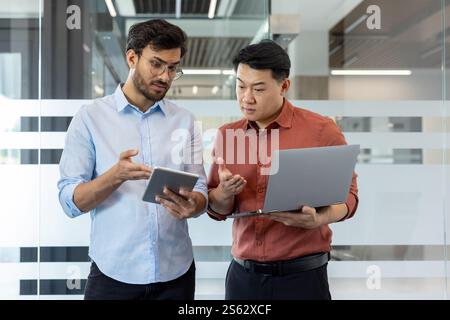 Two diverse office workers discussing work project, serious men in casual clothes standing with tablet computer and laptop, testing new software. Stock Photo