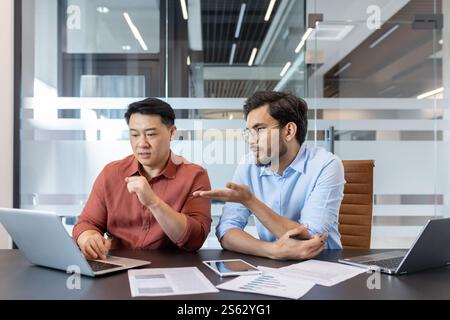 Office workers discussing work project looking at laptop screen, two diverse men sitting at desk with computer, serious and focused in teamwork. Stock Photo