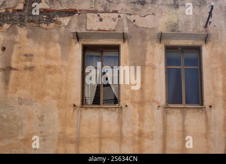 Old weathered orange facade of a house, partly broken facade. Two closed windows, one with curtains. Street in Cefalu, Sicily, Italy. Stock Photo