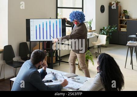 Smiling male boss in clown wig pointing at graphic data on interactive board while explaining it to intercultural subordinates Stock Photo