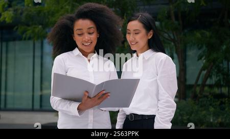 Two African American Asian Korean women females colleagues businesswomen diverse professional coworkers business talking discuss looking paper Stock Photo
