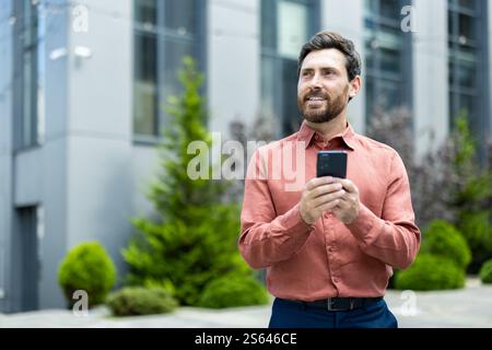 Cheerful smiling man holding phone in hands, thoughtful businessman looking to the side while walking around the city in casual business shirt. Stock Photo