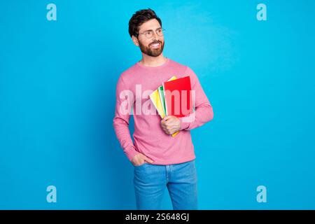 Confident man wearing glasses and casual attire holding colorful notebooks against a vibrant blue backdrop Stock Photo