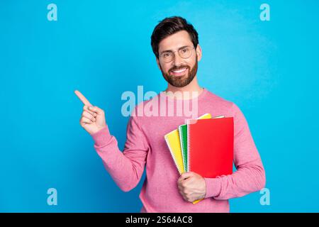 Confident young man in glasses showing direction while holding colorful notebooks against blue background Stock Photo