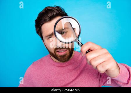 Curious bearded man examining through magnifying glass over blue background in casual wear Stock Photo