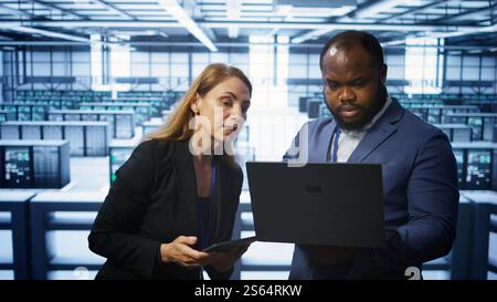 System administrators in data center overseeing server operations using notebook and tablet. Colleagues brainstorming in server hub, ensuring optimal performance and data integrity, camera B Stock Photo