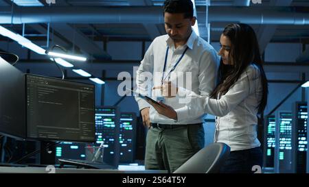 Team of programmers in high tech data center troubleshooting issues using tablet. Teamworking colleagues in server farm using device, establishing performance benchmarks, camera A Stock Photo