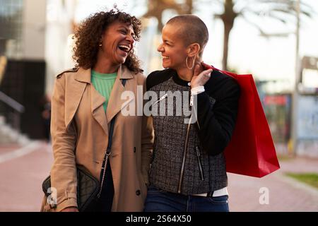 Two multiethnic girlfriends having fun, shopping and laughing while walking on the city street. Stock Photo
