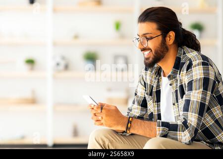 Positive long-haired middle-eastern guy sitting on couch, using cellphone Stock Photo