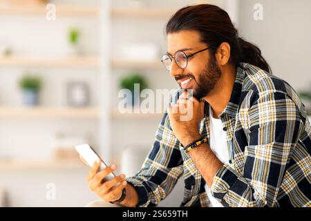 Cheerful long-haired middle-eastern guy hipster sitting on couch, using smartphone Stock Photo