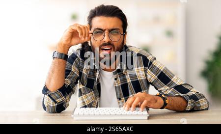 Confused middle-eastern man programmer sitting at worktable, touching glasses Stock Photo