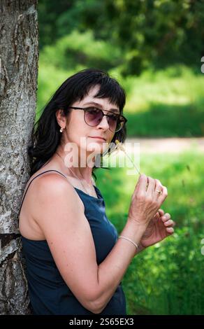 Russian woman, 50 years old, leaning against a birch tree, holding a blade of young grass in a green park during spring. She is wearing sunglasses and Stock Photo