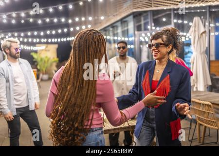 Two multi-ethnic friends women dancing together next to other friends at night party outdoors Stock Photo
