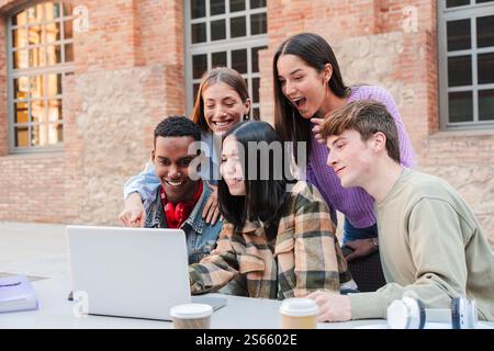 Group of enthusiastic students gathered around a laptop, engaged in collaborative study session, sharing ideas and excitement, representing teamwork and learning among teenagers at university campus. High quality photo Stock Photo