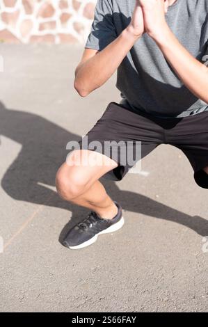 Person doing squats outdoors, legs bent, hands clasped, wearing athletic attire. Stock Photo