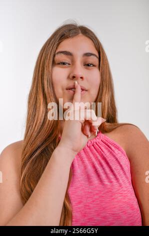 Young woman with long brown hair and pink dress making a silence gesture with her finger on her lips on a white background Stock Photo