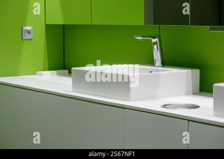 A white, square basin with a chrome mixer, mounted on a white worktop in a bathroom with a bright green tiled wall. Stock Photo