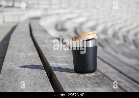 A black thermos with a bamboo lid placed on a wooden bench in a stadium with empty stands in the background. Stock Photo