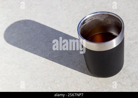 A black thermos with a stainless steel rim, filled with a drink and casting a long shadow on a light surface. Stock Photo