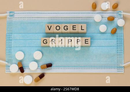 Vogelgrippe - avian influenza in German, spelled with wooden cubes on a surgical mask with scattered pills around. Stock Photo