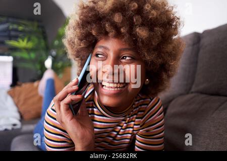 Young African woman at home lying on sofa talking on mobile phone and smiling. Stock Photo