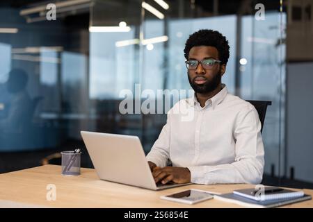 Portrait of a young serious man inside the office with a laptop. Businessman looking at the camera, working in the evening at the workplace. Stock Photo