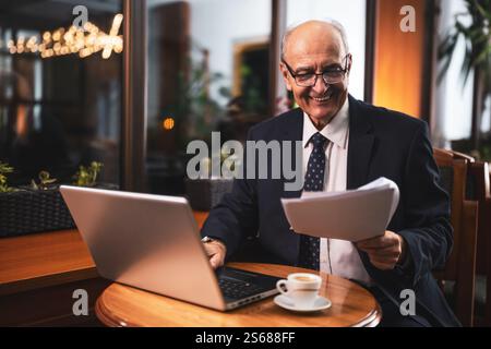 Senior businessman with eyeglasses in elegant suit using laptop and analyzing documents while working and drinking coffee in the city cafe. Stock Photo