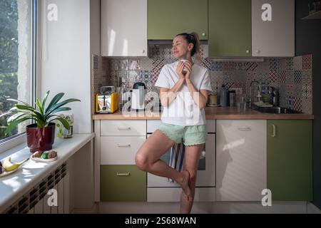 Peaceful woman standing in kitchen, holding cup of hot beverage and dreamy looks out of window. Stock Photo