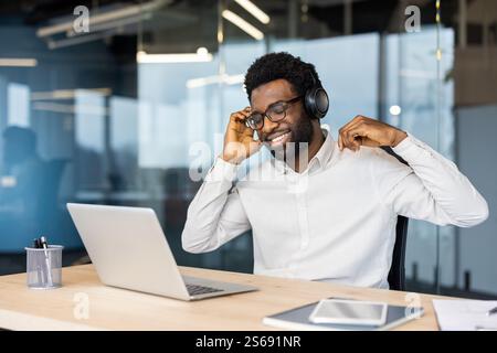 Young man at workplace working with laptop, listening to music, smiling and singing along. Worker in headphones happily resting and relaxing while working in the evening. Stock Photo