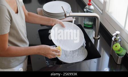 Woman washing and rinsing dishes by hand under a stream of water in the kitchen sink. Washing dishes process. Stock Photo