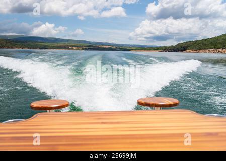View from the back of a boat moving through Furnas Dam Lake, leaving behind a foamy wake. There's a wooden table with two wooden stools. Stock Photo