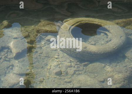 An old tire and other waste have been discarded into the small water stream that flows through the town with the tire now overgrown with algae. Stock Photo