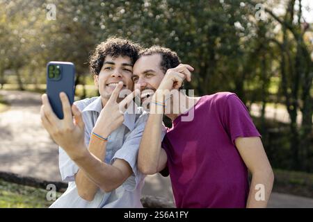 Happy brazilian gay couple wearing rainbow bracelets taking selfie with smartphone and making victory sign in park Stock Photo