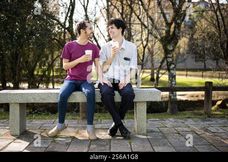 Two men sitting on a bench in a park, drinking coffee and chatting Stock Photo