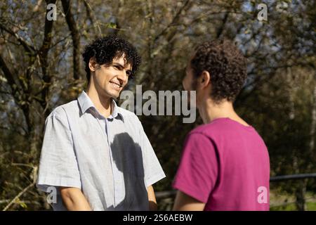 A gay couple looks at each other tenderly while holding hands, showing their love in a sunny park Stock Photo