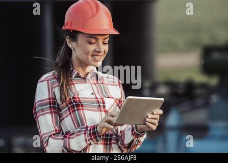 Portrait of pretty young female engineer with helmet standing in front of irrigation system plant in agricultural field and working on tablet Stock Photo