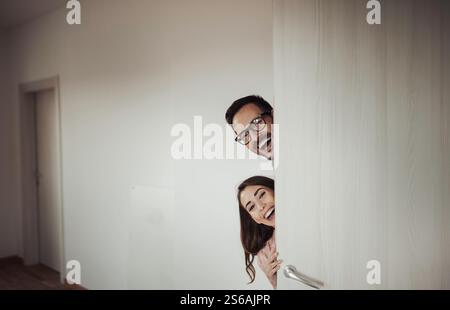 Excited young couple peeking behind open door and welcoming guests in new empty apartment Stock Photo