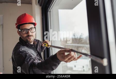 Portrait of young construction worker measuring window dimensions with tape during renovation in apartment Stock Photo