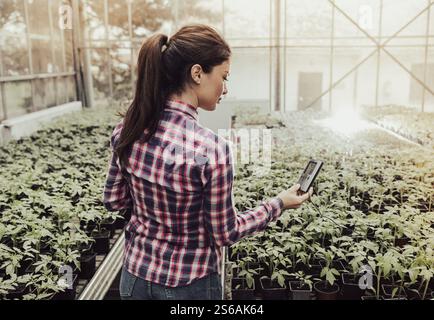 Pretty young farmer woman checking temperature in greenhouse with vegetable seedlings in flower pots Stock Photo