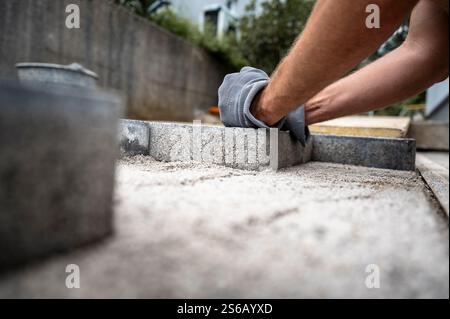 Closeup of a person wearing gloves carefully placing concrete pavers on a sandy base during a patio construction project. Stock Photo