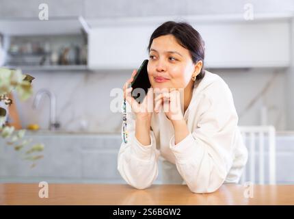 Young woman leaning on table in kitchen, taking on smartphone Stock Photo