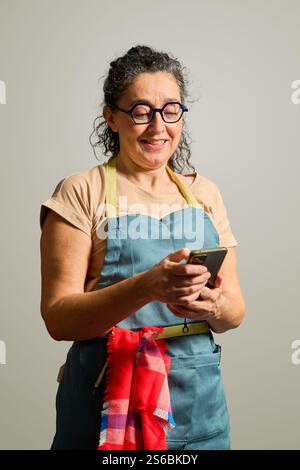 Happy mature woman chef wearing an apron and glasses, smiling while using her smartphone to browse recipes and connect online Stock Photo