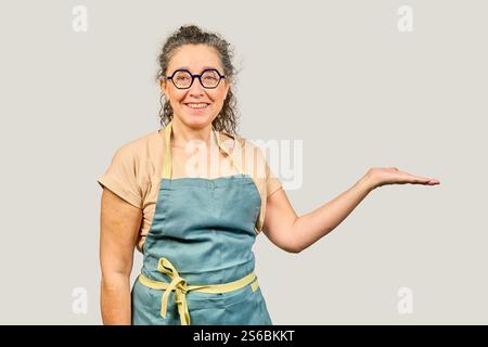 Friendly mature woman wearing apron and glasses is showing something on the palm of her hand, isolated on gray background Stock Photo