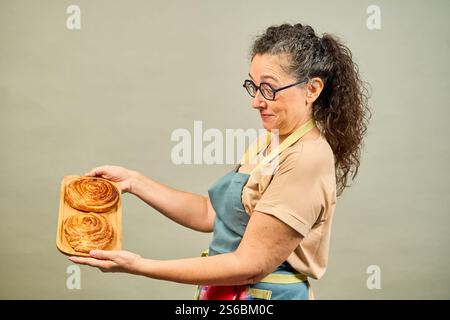 A woman is holding two cinnamon rolls in her hands. She is wearing an apron and glasses Stock Photo