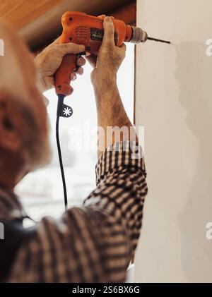 Senior craftsman drilling hole in wall beside window with electrical screwdriver indoors Stock Photo