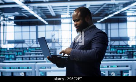 Computer scientist in server hub checking recovery plan on laptop, monitoring hardware energy consumption. IT specialist uses notebook in data center facility, ensuring rigs are functioning, camera A Stock Photo