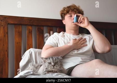 Young man sitting on bed with a cat and using inhailer for asthma attack Stock Photo