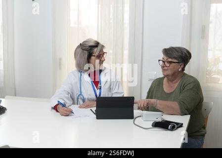 Female doctor taking notes while visiting a senior patient at home and using a tablet with a blood pressure monitor on the table Stock Photo