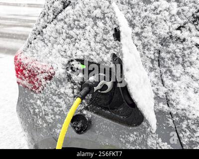 Charging an electric vehicle in winter. A yellow charging cable is connected to the car's charging socket while snow has accumulated on the bodywork, Stock Photo