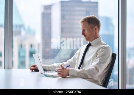 Young busy businessman looking at computer writing notes at work desk in office. Stock Photo
